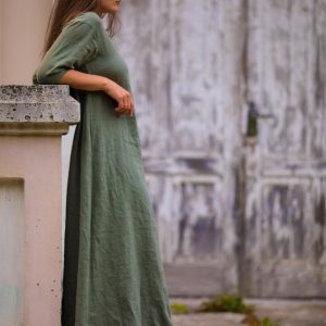 Creek linen dress by Knock Knock Linen in sage green—one loose maxi trapeze tunic with round neck and elbow-length sleeves. Shown on model standing in sandals on pavement by stone steps, weathered door behind. Linen summer dress with sleeves.