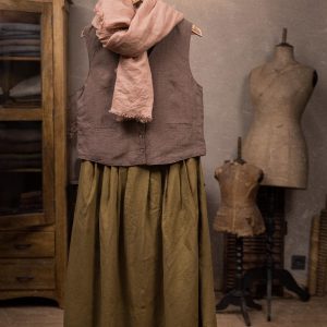 Olive green linen maxi skirt displayed full-length on a hanger, front view, with a gathered waist, deep vertical folds, wide hem, brown sleeveless vest above, and pale pink scarf at the neck.