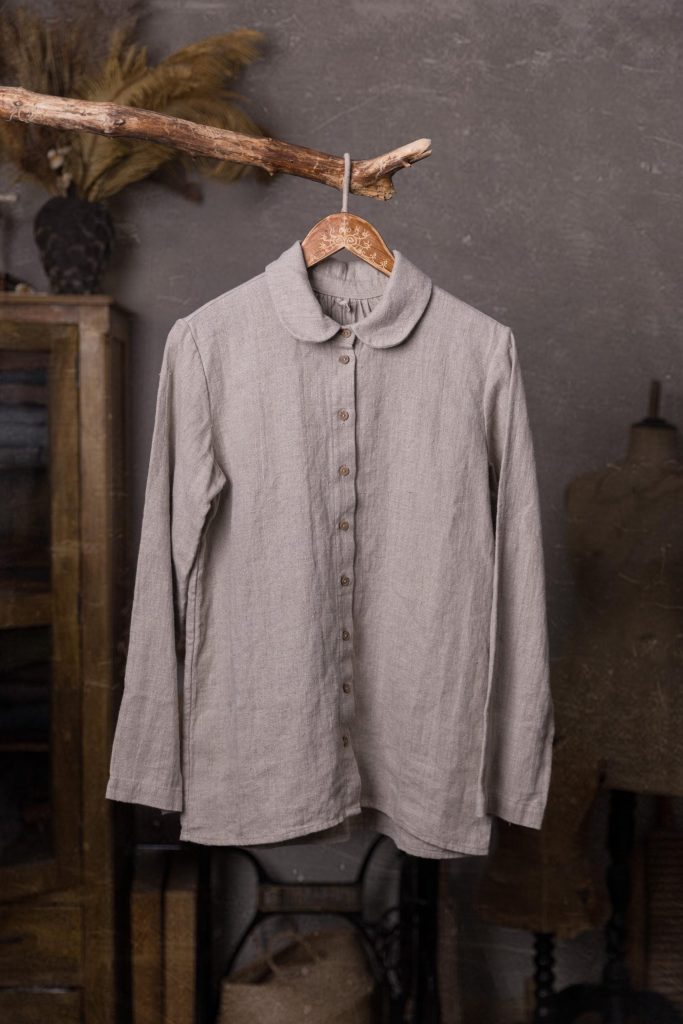 Light gray linen shirt shown full-length from the front on a wooden hanger, with a rounded collar, long sleeves, a narrow button placket, small round buttons, visible creases and a gently curved hem.